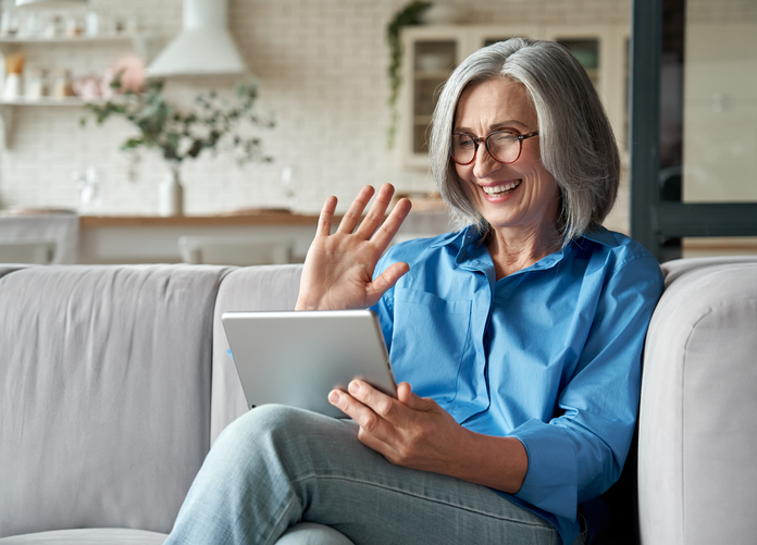 Happy 60s older mature middle aged adult woman waving hand holding digital tablet computer video conference calling by social distance virtual family online chat meeting sitting on couch at home. TELEMEDICINE WITH DERMATOLOGISTS OF BIRMINGHAM